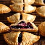 Golden blueberry hand pies on a cooling rack with one cut open showing filling