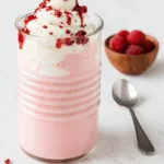 A tall glass of creamy raspberry milkshake topped with whipped cream, raspberry coulis, and freeze-dried raspberry crumbs, served with fresh raspberries on the side.